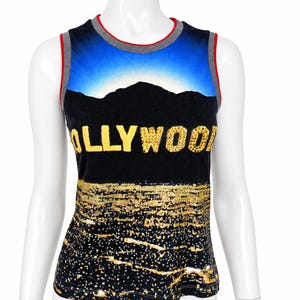 May include: A sleeveless tank top featuring a black background with a graphic print of a mountain silhouette against a blue sky. The word OLLYWOOD is printed in large gold letters across the chest, and the lower portion features a gold glitter pattern.