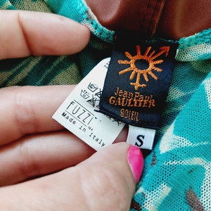 May include: A turquoise and brown patterned mesh top with a black tag that says "Jean Paul Gaultier Soleil" and a white tag that says "Fuzzi S.r.l. Made in Italy" and a white tag with the letter "S" on it.