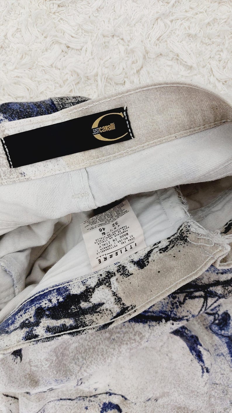 May include: A white and blue patterned pair of jeans with a black label that says "Just Cavalli" and a white tag with the text "T.T.I.L.I.E.R.E.  CONA  P.T. TORNARE  MADE IN ITALY  D.I.  BOL.128  49  32  F.  4  PANTALONI  LAVAGGIO A FREDDO  NON CANDEGGIARE  STIRARE A BASSA TEMPERATURA  NON ASCIUGARE IN TAMBURO  LAVAGGIO A SECCO  CON PERCLORETIEN  "