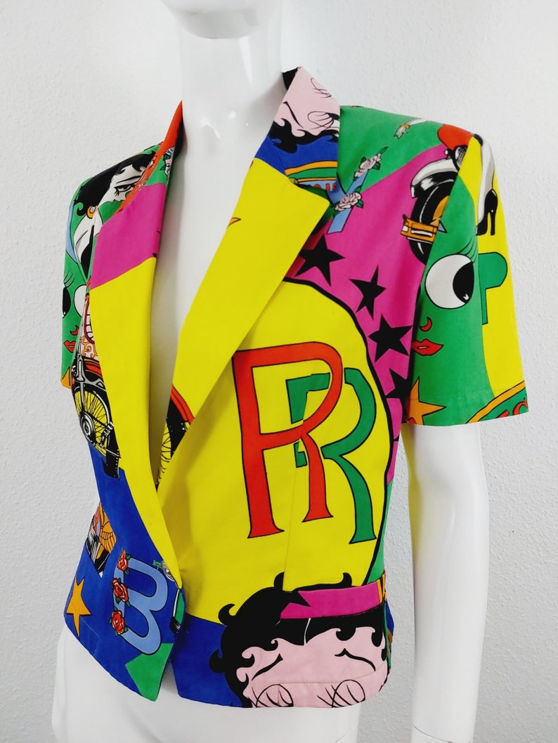 May include: A short-sleeved, multi-coloured blazer with a patchwork design. The blazer features a large "PR" logo in red and green letters on a yellow background. The blazer also features a cartoon character with black hair and a pink dress.