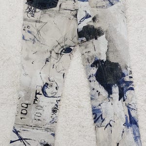 May include: White denim jeans with a blue and black abstract print. The jeans have a brown leather patch with a letter C on the back waistband.