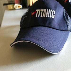 May include: A navy blue baseball cap with a white mesh back and a white embroidered "TITANIC" logo. A small red and white flag is embroidered on the front of the cap.