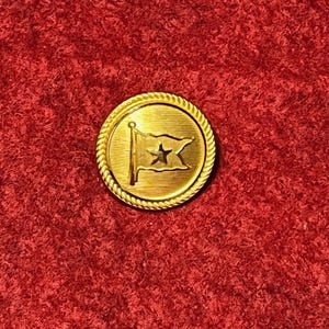 Puede incluir: Un botón redondo dorado con un borde en forma de cuerda. El botón presenta un diseño de bandera con una estrella en el centro. El botón está sobre un fondo rojo.