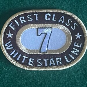 Może przedstawiać: Owalna naszywka w kolorze brązowym i złotym z białym haftem. Naszywka zawiera napis "First Class White Star Line" i ma białą cyfrę "7" w środku.