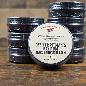 May include: A collection of Officer Pitman's Bay Rum beard and mustache balm jars. The product is packaged in small, black metal tins with a white label. The label includes the text "OFFICIAL GROOMING SUPPLIES" and "Net Wt. 2 oz (56 g)".