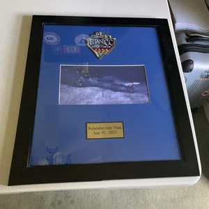 Puede incluir: Una foto enmarcada de una imagen en blanco y negro del naufragio del Titanic con una placa dorada que dice "Remember the Titan June 18, 2023". El marco es negro y tiene un paspartú azul. Hay un parche dorado en el paspartú que dice "Titanic Survey Expedition 2023".