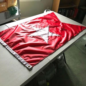 Peut inclure: Un drapeau rouge avec une étoile blanche au centre. L'étoile est bordée de coutures blanches. Le drapeau est fait d'un matériau brillant et soyeux.