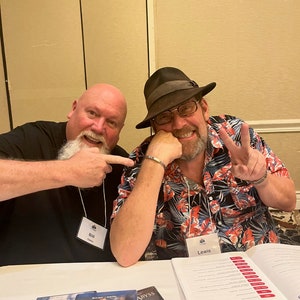 May include: Two men are sitting at a table with a white tablecloth. The man on the left is wearing a black shirt and has a white beard. He is pointing to his name tag, which says "Bill". The man on the right is wearing a black and red Hawaiian shirt, a black hat, and glasses. He is holding up two fingers in a peace sign. His name tag says "Lewis". There are three DVD cases on the table, one for "Titanic", one for "The Abyss", and one for "Titanic".