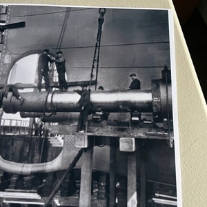 Pu&ograve; includere: Foto in bianco e nero di un grande tubo di metallo che viene sollevato da una gru. Diversi operai sono in piedi su una piattaforma attorno al tubo.