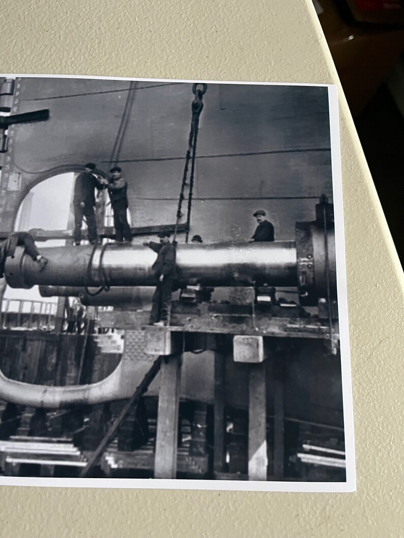 Pu&ograve; includere: Foto in bianco e nero di un grande tubo di metallo che viene sollevato da una gru. Diversi operai sono in piedi su una piattaforma attorno al tubo.