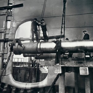 Pu&ograve; includere: Foto in bianco e nero di un grande cannone di metallo che viene sollevato da una gru. Diversi uomini stanno lavorando al cannone, che sta per essere installato su una nave.