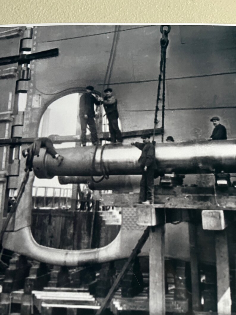 Pu&ograve; includere: Foto in bianco e nero di un grande cannone di metallo che viene sollevato da una gru. Diversi uomini stanno lavorando al cannone, che sta per essere installato su una nave.