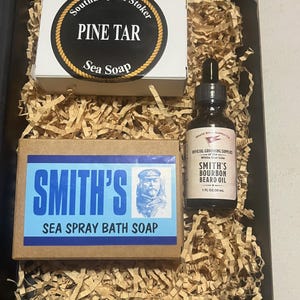 Może przedstawiać: Zestaw upominkowy zawierający kostkę mydła Pine Tar Sea Soap, kostkę mydła Smith's Sea Spray Bath Soap oraz butelkę olejku do brody Smith's Bourbon. Artykuły są zapakowane w czarne pudełko z pociętym papierem.