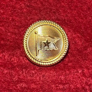 May include: A gold-colored, round button with a detailed design. The button features a flag with a star and a rope-like border. The background is a textured red fabric.