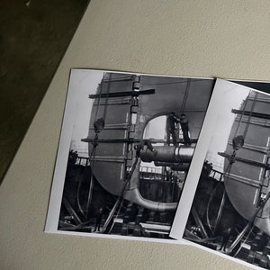Pu&ograve; includere: Fotografia in bianco e nero di una grande struttura metallica in fase di assemblaggio da parte di due operai. La struttura &egrave; sollevata da una gru e sostenuta da travi in legno. La fotografia &egrave; scattata da un angolo basso, guardando verso l'alto la struttura.