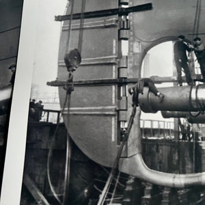 Pu&ograve; includere: Foto in bianco e nero di una grande parte di nave in metallo che viene sollevata da una gru. La parte viene abbassata in un bacino di carenaggio. I lavoratori sono visibili nella foto.