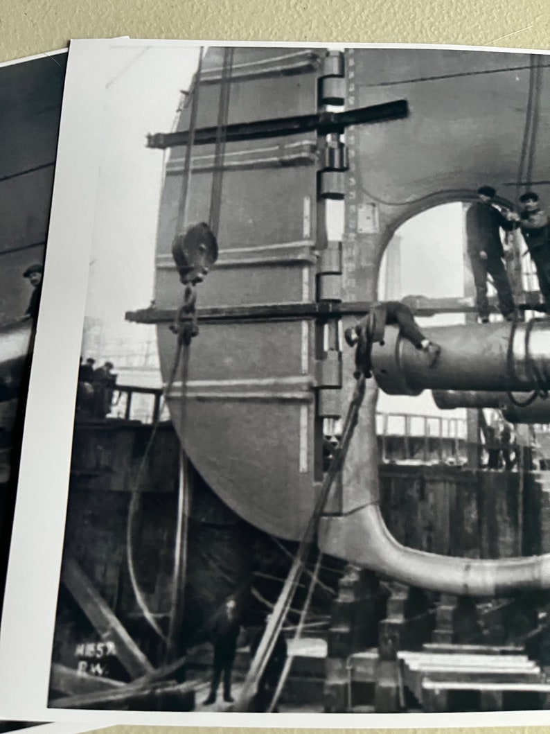 Pu&ograve; includere: Foto in bianco e nero di una grande parte di nave in metallo che viene sollevata da una gru. La parte viene abbassata in un bacino di carenaggio. I lavoratori sono visibili nella foto.