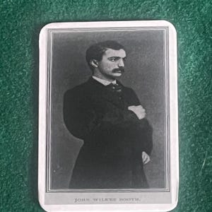 Op de afbeelding: Zwart-wit portret van een man in een donker pak, armen gekruist, met de tekst "JOHN WILKES BOOTH" onderaan. De afbeelding staat op een rechthoekige kaart met afgeronde hoeken.