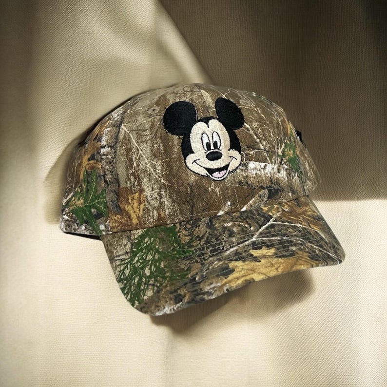 May include: A camouflage baseball cap with an embroidered Mickey Mouse face. The cap has a curved peak and is made of fabric with a brown, green, and tan pattern. The Mickey Mouse design is black, white, and pink.