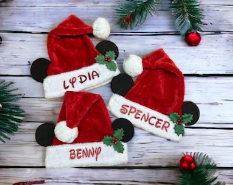 May include: Three red Santa hats with white trim and pom-poms, each featuring a name in glitter lettering: Lydia, Spencer, and Benny. Black mouse ears and holly accents complete the festive design, perfect for Christmas.