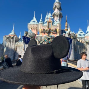 May include: A black fedora hat with Mickey Mouse ears attached to the sides. The hat is being held up in front of a large, ornate castle.