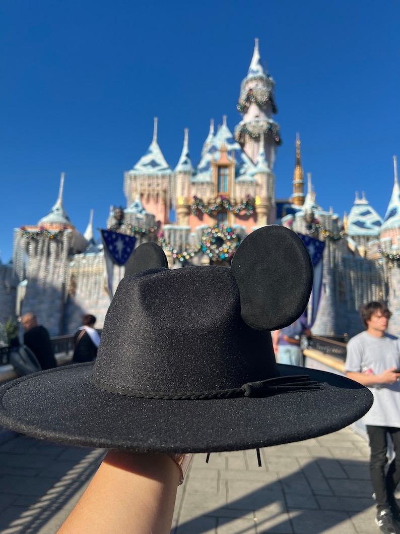 May include: A black fedora hat with Mickey Mouse ears attached to the sides. The hat is being held up in front of a large, ornate castle.