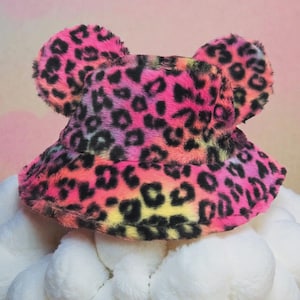 May include: A vibrant bucket hat with a pink, orange, yellow, and purple leopard print pattern. The hat features two large, fluffy ear-like accents on top. The hat is made of a soft, plush material.