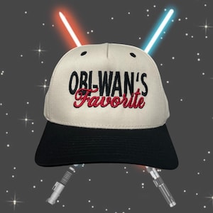May include: Cream-colored baseball cap with a black brim. The cap features the embroidered text "Obi-Wan's Favorite" in black and red. The background includes lightsabers and stars.