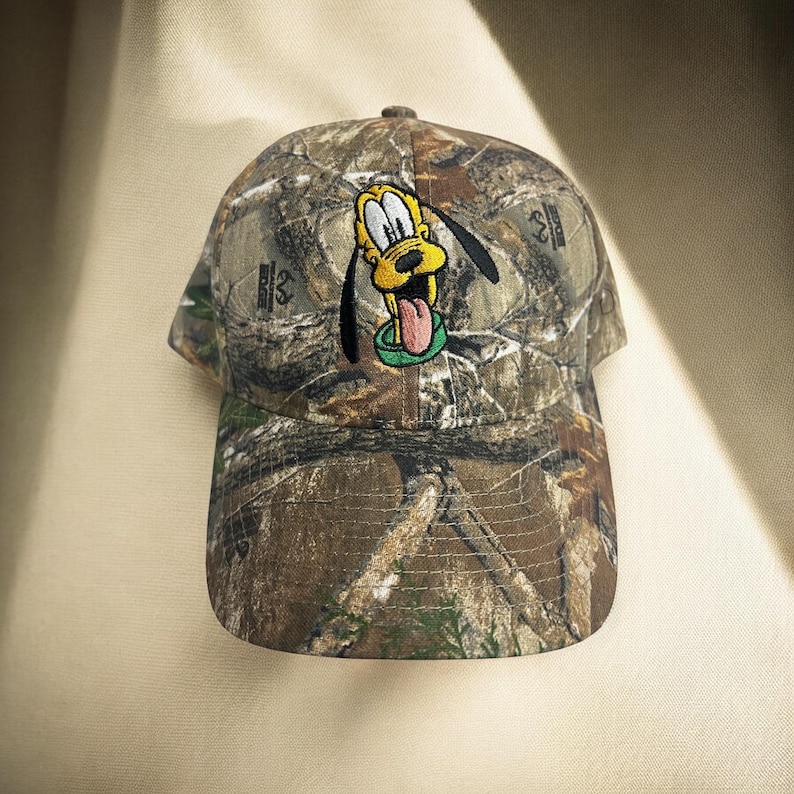 May include: A camouflage baseball cap with an embroidered Pluto design. The cap has a curved peak and a six-panel design. The Pluto design is in yellow, black, and red, with a green collar. The cap is made of camouflage fabric.