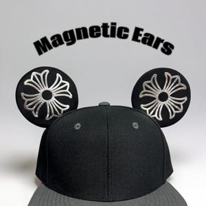 May include: Black baseball cap with two round, black magnetic ears. Each ear features a silver floral design. The words "Magnetic Ears" are printed above the cap in black letters.