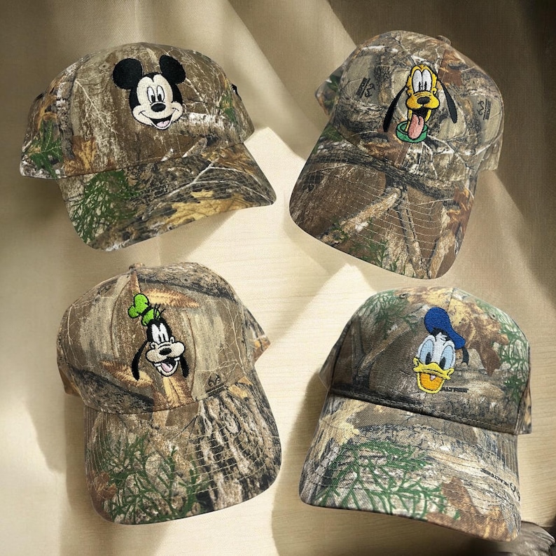 May include: Four camouflage baseball caps with embroidered Disney characters: Mickey Mouse, Pluto, Goofy, and Donald Duck. Each cap has a curved peak and an adjustable strap. The caps are in a woodland camouflage pattern.