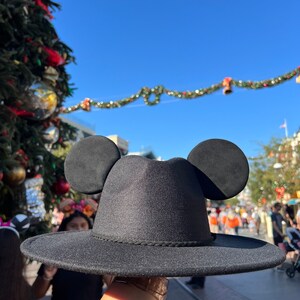 May include: A black fedora hat with Mickey Mouse ears. The hat has a wide brim and a black band around the crown.