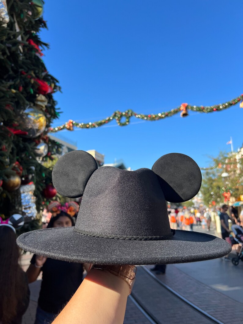May include: A black fedora hat with Mickey Mouse ears. The hat has a wide brim and a black band around the crown.