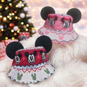 May include: Two red and pink bucket hats with Mickey Mouse ears and a Christmas-themed pattern. The hats feature a repeating pattern of Mickey Mouse's face and a festive design.