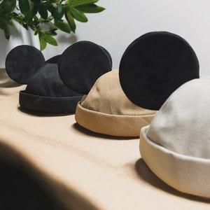 May include: Three different styles of black, beige, and white hats with a Mickey Mouse ear design. The hats are made of a soft, fabric material.