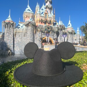May include: A black fedora hat with Mickey Mouse ears, in front of a blurred background of Sleeping Beauty's castle at Disneyland.