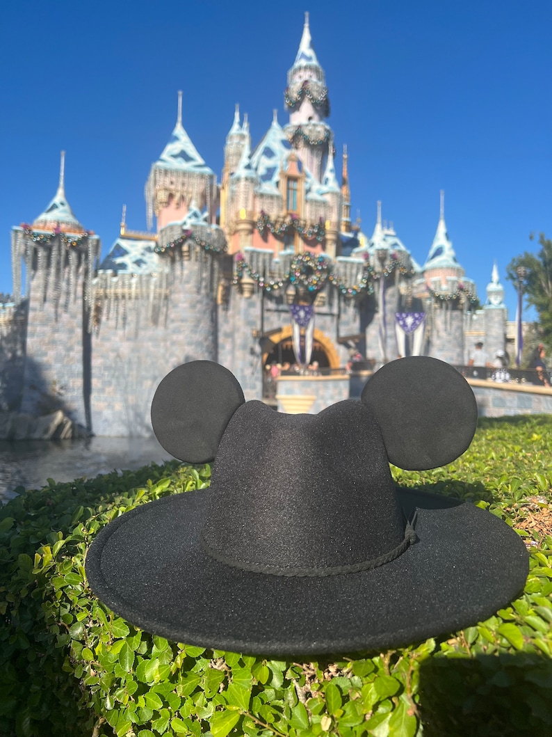 May include: A black fedora hat with Mickey Mouse ears, in front of a blurred background of Sleeping Beauty's castle at Disneyland.