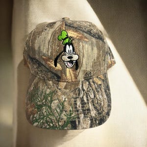 May include: A camouflage baseball cap with an embroidered Goofy design. The cap has a curved peak and a classic six-panel construction. The hat's pattern incorporates shades of brown, green, and tan.