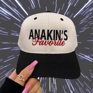May include: A baseball cap with a black brim and a cream-colored crown. The text "ANAKIN'S Favorite" is embroidered on the front in black and red. The cap has a classic six-panel design with a curved brim.