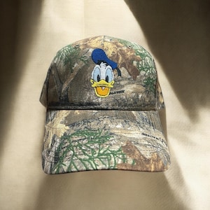 May include: A camouflage baseball cap with an embroidered Donald Duck design. The cap has a curved peak and a classic six-panel construction. The Donald Duck image is in blue, yellow, and white.