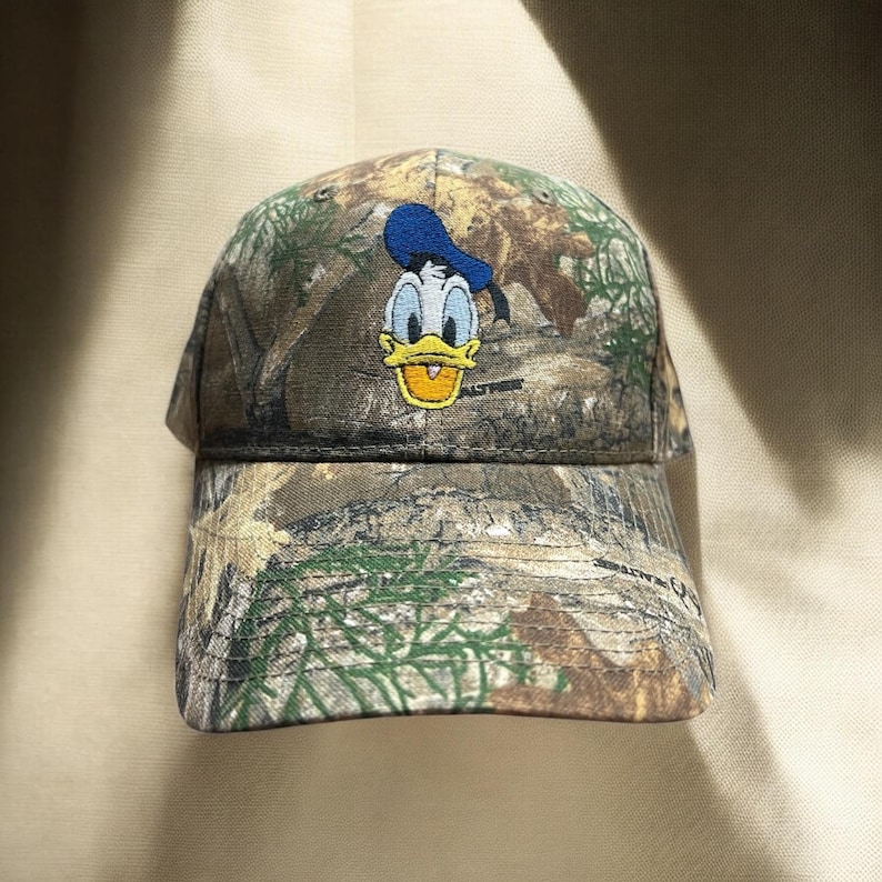 May include: A camouflage baseball cap with an embroidered Donald Duck design. The cap has a curved peak and a classic six-panel construction. The Donald Duck image is in blue, yellow, and white.