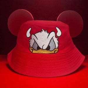 May include: Red bucket hat with attached Mickey Mouse ears. The hat features an embroidered image of Donald Duck's face in white, gray, and yellow. The hat is made of a soft material and is suitable for casual wear.