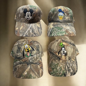 May include: Four camouflage baseball caps featuring Disney characters Mickey Mouse, Donald Duck, Pluto, and Goofy. Each cap has an embroidered character design on the front and a curved brim. The caps are in shades of brown, green, and tan.