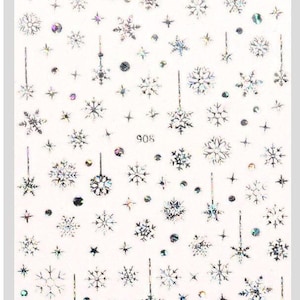 May include: A sheet of white nail stickers with a variety of silver snowflake designs. The sheet includes a number of different snowflake shapes and sizes, as well as some small stars and dots. The sheet is labeled "908".