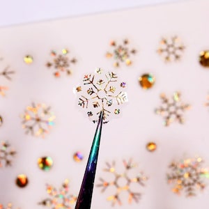 May include: A close-up of a gold snowflake sticker being held by tweezers. The sticker is iridescent and has a holographic finish. The sticker is surrounded by other gold snowflake stickers on a white background.