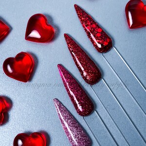 Puede incluir: Cinco tonos diferentes de esmalte de uñas de purpurina roja en palitos de exhibición sobre un fondo gris. Los colores del esmalte de uñas van de un rosa claro a un rojo intenso. Corazones rojos están dispersos alrededor del esmalte de uñas.