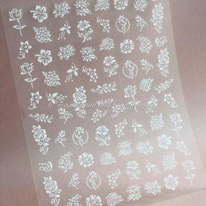 Könnte beinhalten: Blatt mit weißen Nagelaufklebern mit verschiedenen floralen Designs. Das Blatt enthält Rosen, Tulpen und andere Blumen.