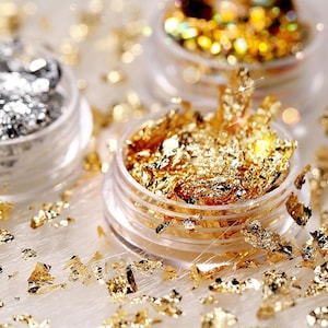 May include: Three small jars filled with gold and silver leaf flakes. The jars are clear plastic with silver lids. The flakes are scattered around the jars on a white surface.