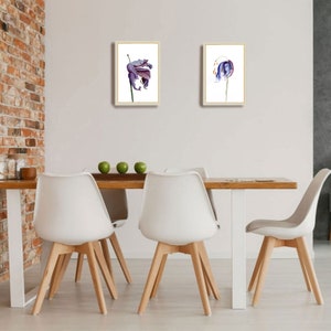 May include: Two framed floral art prints. Each print features a watercolor painting of a purple and blue flower on a white background. The frames are a light wood color. The prints are displayed above a wooden table with white chairs.