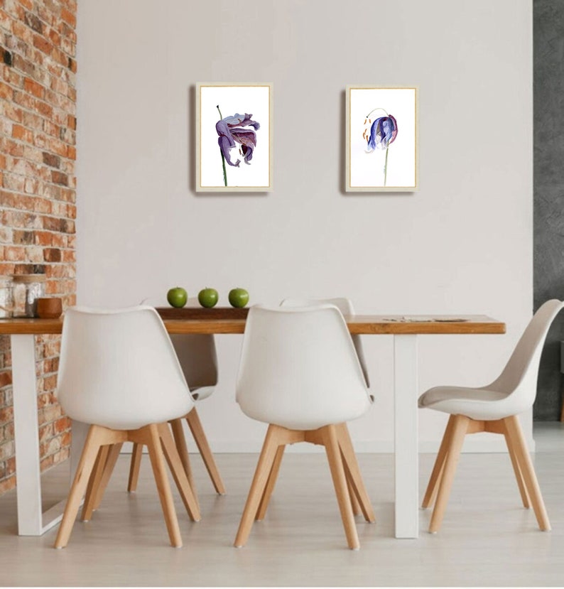 May include: Two framed floral art prints. Each print features a watercolor painting of a purple and blue flower on a white background. The frames are a light wood color. The prints are displayed above a wooden table with white chairs.